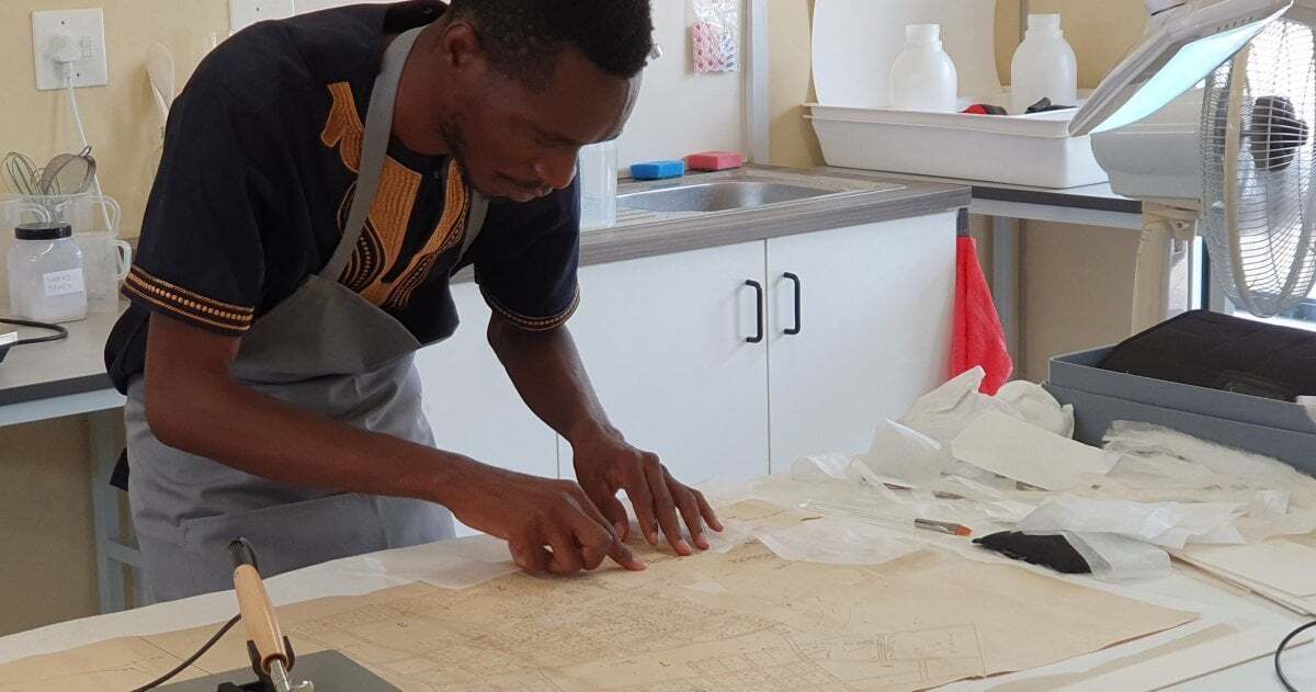 Ein junger Mann aus Namibia steht an einem Tisch und bessert Risse eines alten Gebäudegrundrisses aus.