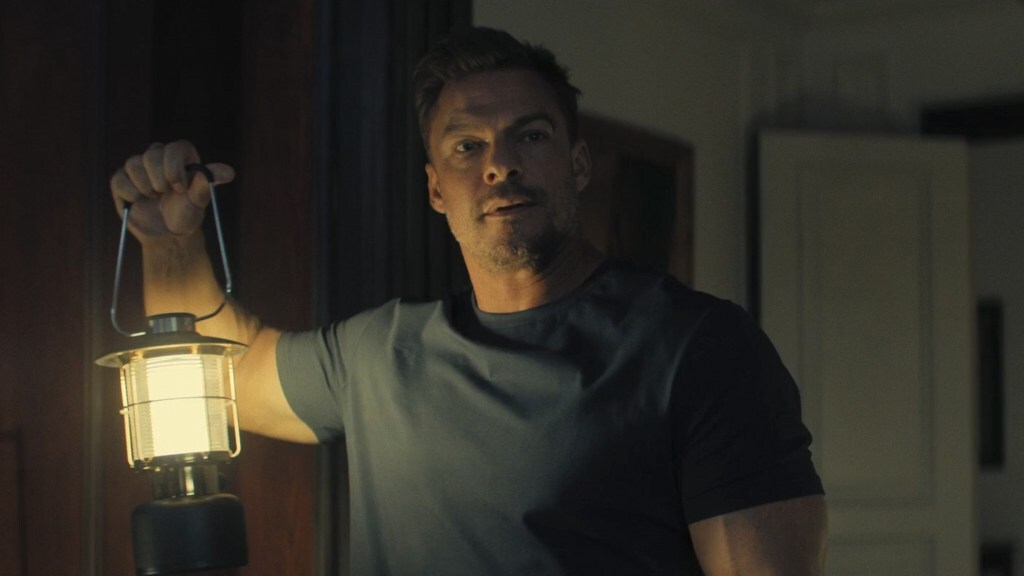 Alan Ritchson holds up a lantern in a dark room in a still from 'Reacher'.