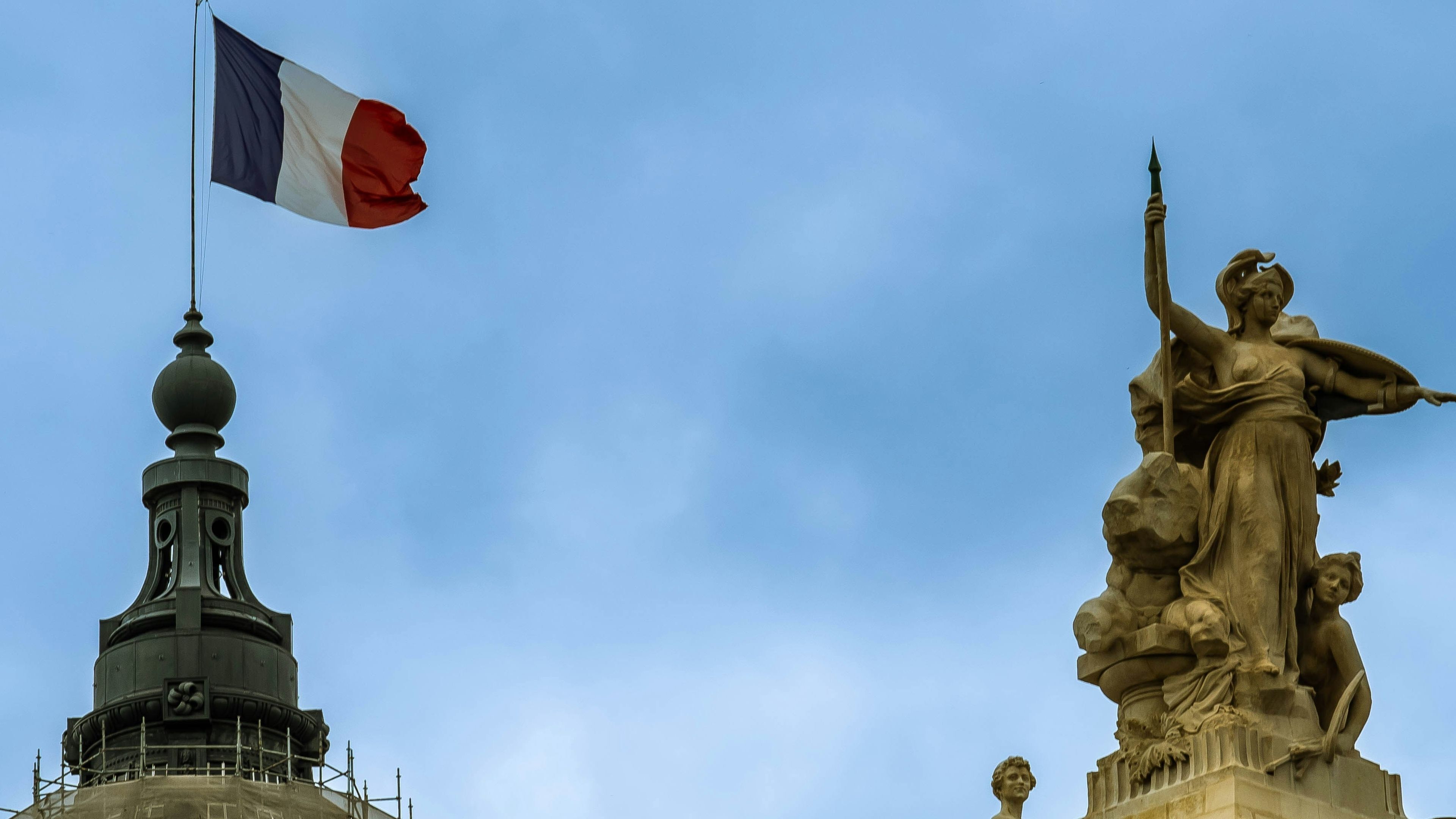 A French flag flutters atop a building (Pourya Gohari/Unsplash)