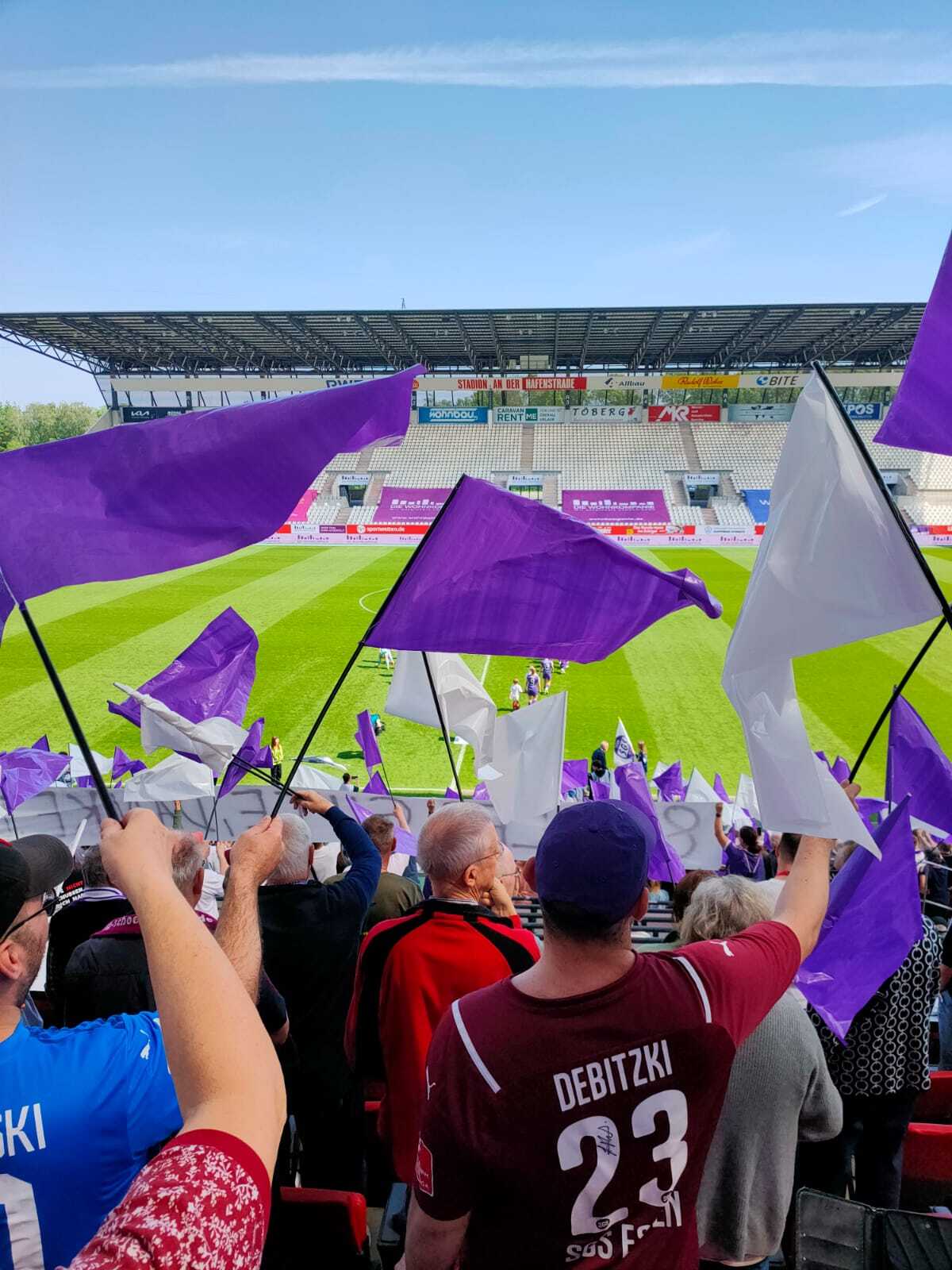 Blick ins Stadion an der Hafenstraße von der Haupttribüne Höhe Mittellinie aus dem Fanblock heraus. Fans in SGS Trikots schwenken lilane und weiße Fahnen, im Hintergrund laufen die beiden Teams gerade vor Anpfiff ins Stadion ein.