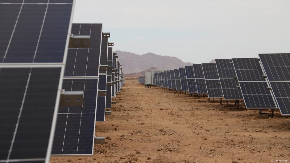Schräg aufgestellte Solarpaneele in langen Reihen vor Bergen auf kargem Boden