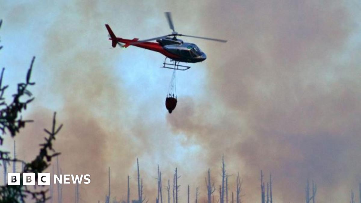 Photo of a bright orange blaze lighting up a hazy sky. Smoke can be seen rising from the hills, as well as outlined trees. A helicopter can be seen flying over the trees, with a water bucket attached to the helicopter. 