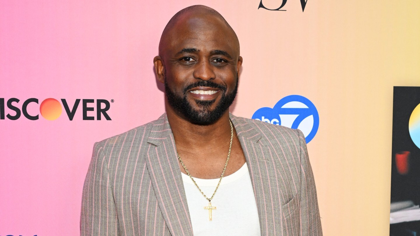 Wayne Brady poses at an event in a tan suit and white t-shirt.