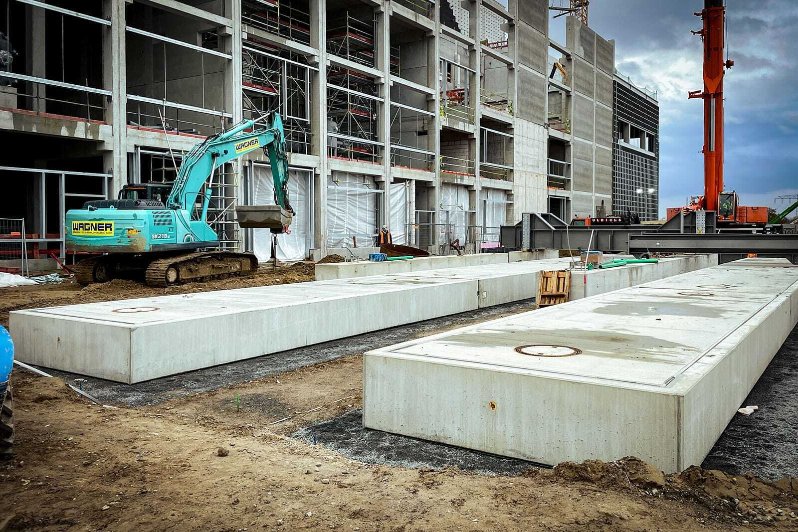 Grüner Bagger neben zwei 30 Meter langen Unterflur-Fahrzeugwaagen aus Beton auf der Baustelle des neuen Klärwerks Waßmannsdorf.