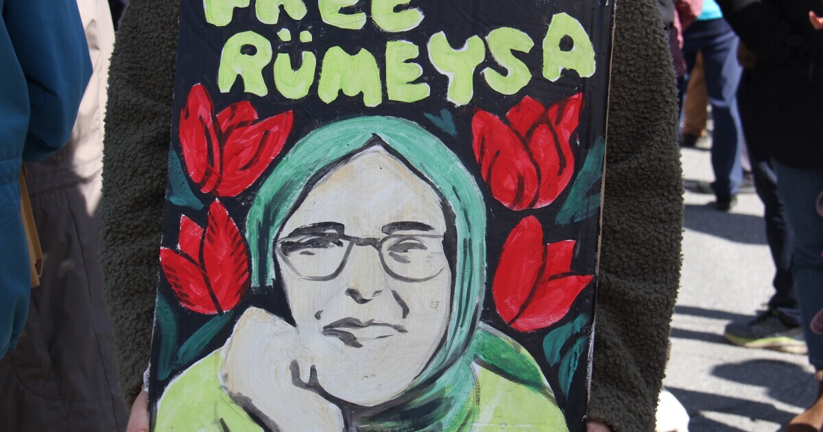 A painted sign shows a woman wearing glasses and wearing a head covering with the words "free Rümeysa" 