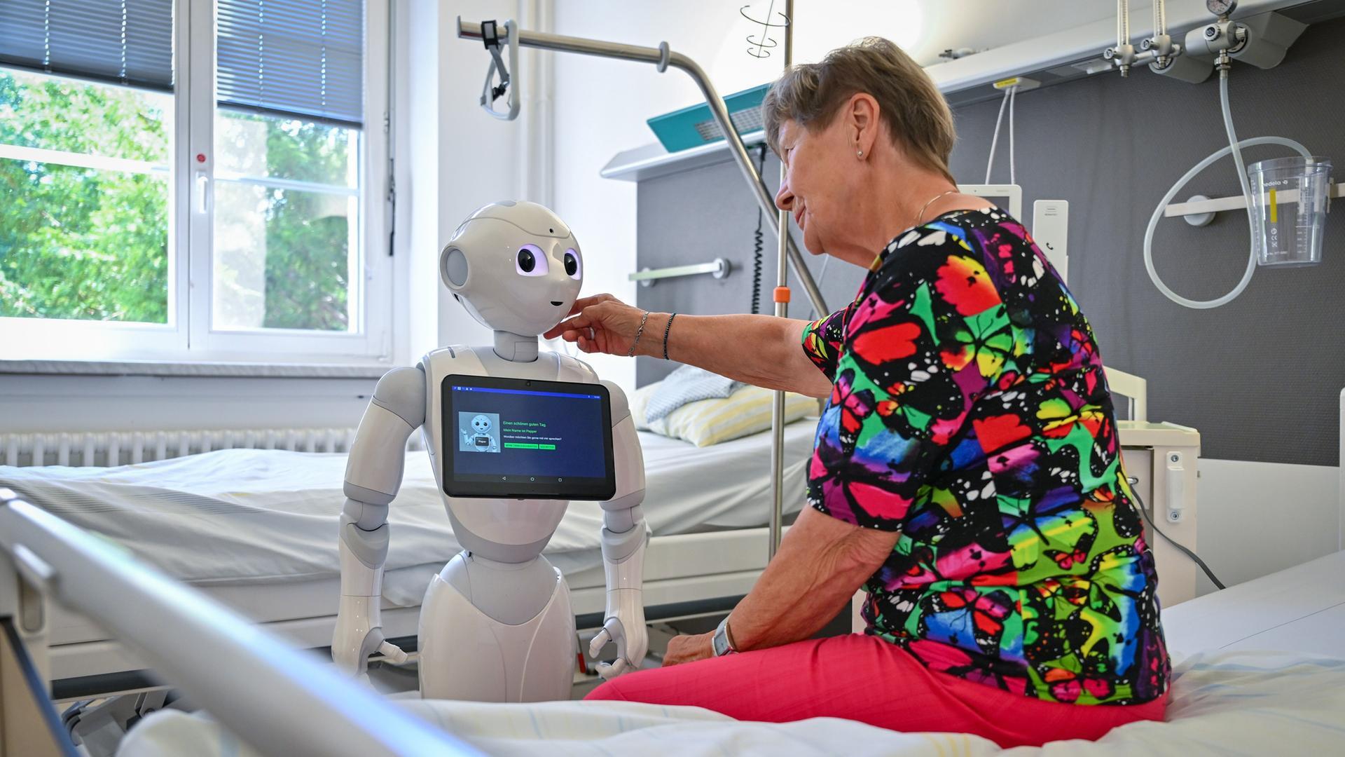 Eine ältere Dame sitzt auf einem Pflegebett und streicht einem humanoiden Roboter, der vor ihr steht, mit der rechten Hand über die Wange.