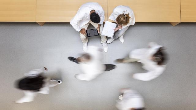 Des personnes en sarrau marchent dans un corridor; d'autres personnes sont assises et regardent une tablette et un cahier.