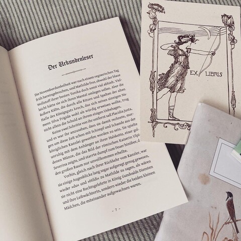 Ein offenes Buch das den Beginn eines neuen Kapitels zeigt. Die Überschrift, ist passend zu den Pergamenten auf dem Cover, in Fraktur gesetzt. Daneben liegt ein Notizbuch und eine Postkarte mit Ex Libris Aufdruck. 