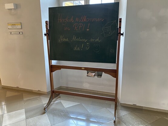 Man sieht eine sehr alte Tafel in einem Gang. Auf der Tafel steht:
Herzlich willkommen im RPI! Neue Medien sind da!