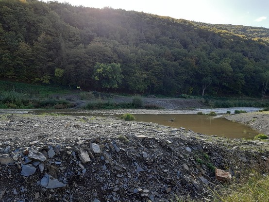 Im Vordergrund Schutt und Geröll. Wasserfläche und der Fluss. Auf der anderen Seite ein ansteigender Weg und ein bewaldeter Hügel. 