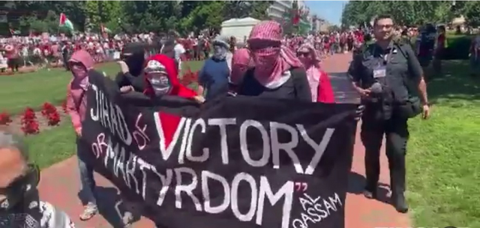 Mit Kaffia verhüllte „Demonstranten“ tragen einen schwarzen Banner mit den Worten „Jihad of Victory or Martyrdom - Al Qassam“ vor sich her. Das V von Victory ist rot ausgemalt. Ein Polizist läuft dahinter mit.