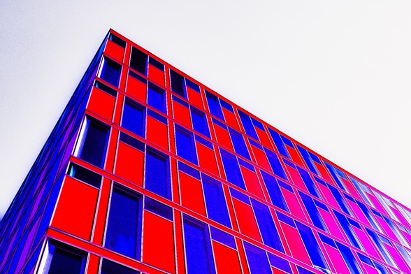 Das Bild zeigt ein Gebäude mit einem modernen architektonischen Design. Die Fassade ist mit roten und blauen Paneelen gestaltet, die ein auffälliges Muster bilden. Die Perspektive ist von einem niedrigen Winkel nach oben gerichtet, was die Höhe und die geometrischen Linien des Gebäudes betont. Das Gebäude steht gegen eine weiße Fläche, die den erwarteten Himmel ersetzt. Die Farben sind digital verstärkt, was dem Bild ein lebendiges und beeindruckendes Aussehen verleiht.