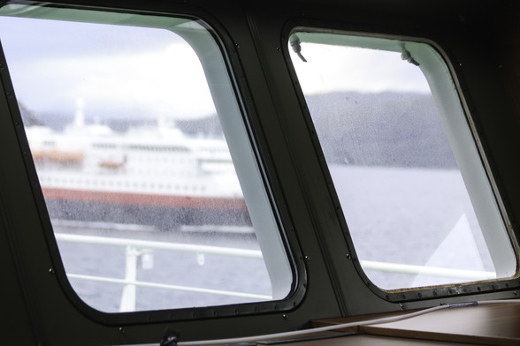 Zwei Fenster auf dem Beobachtungsgang der Polarstern mit Blick nach hinten. Im Hintergrund ein Berg am Ufer eines Fjordes, und parallel zu uns eine schwarz rot weiße Fähre, die uns langsam überholt. Auf den schrägen Fensterscheiben sitzt ein feiner Film aus Gischt. 