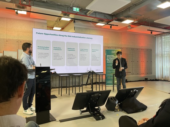A person in a light blue shirt stands by a podium, and another person in a dark suit speaks into a microphone. They are presenting in front of a large screen displaying text about future opportunities in RAG industrialization. An audience member is visible in
