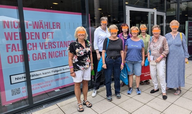 Gruppenbild von 8 Frauen. 3 Frauen im Hintergrund und 4 Frauen vorne.
Sie stehen vor einem Gebäude mit einer großen Glasfront. Der Platz auf dem sie stehen hat Bodenfliesen.
Hinter dem Fenster ist ein Überlebensgroßes Plakat auf dem steht: 