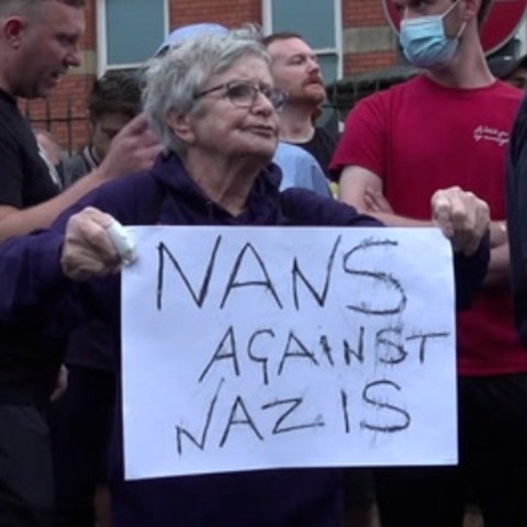 Eine alte Dame in einer Gruppe stehend hält eine beschriebene Pappe in der Hand auf der steht: 