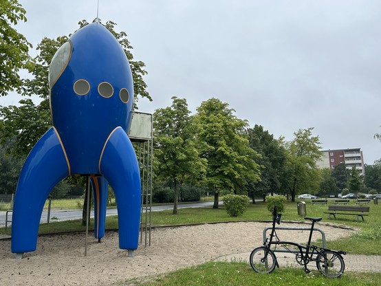 Foto eines Spielplatzes in Bernau, links ist eine blaue Rakete mit Kletterleiter. Es ist die Zwillingsschwester der Fair-Dust vom CCC, 
rechts ein Brompton an einem Fahrradbügel, 
die Rakete steht im Sand,
rundherum ist eine Grünfläche im Hintergrund eine Straße, Bäume und ein 6-Geschoß-Hochhaus