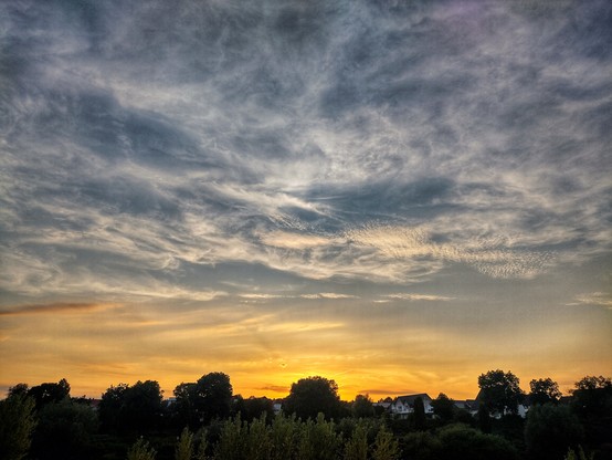 Foto des Himmels, nur am unteren Bildrand sind einige Bäume und Häuser erkennbar. In Richtung Sonnenuntergang fotografiert ist der Himmel unten orange-Rot und geht nach oben in blau mit vielen Schleierwolken über.