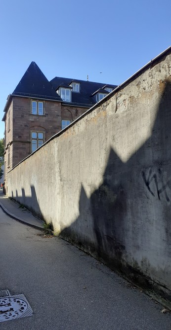 Schmale Gasse, rechts eine durchgehende Mauer, an deren  Ende weiter vorne ein Haus mit kleinen 