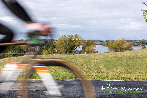 Ein Fahrrad im Vordergrund fährt an einem See vorbei. Das Fahrrad ist schnell und verwischt, man erkennt die Streifen des Fahrrads in blau, orange und gelb.