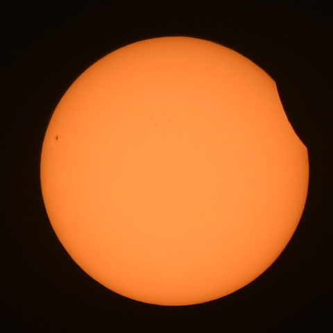 Foto der Sonne durch einen starken Lichtfilter. Am rechten Rand ist ein schmaler Teil der Sonne durch den Mond verdeckt. am linken Rand ein typischer Sonnenfleck.