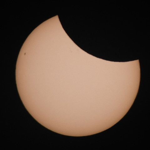 Fotografie der Sonne durch einen starken Lichtfilter. Oben rechts fehlt ein größeres Stück, das durch den Mond verdeckt ist. Links ein kleiner dunkler Sonnenfleck, oberhalb der Mitte und nahe des Mondrandes weitere schwachdunkle Flecken auf der Sonnenoberfläche.