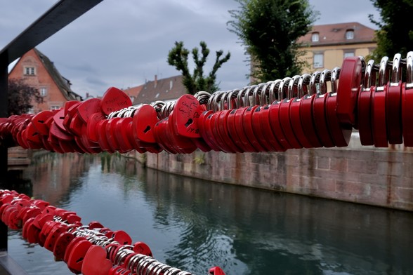Im Hintergrund sieht man alte Gebäude, die an einem Kanal stehen. Im Vordergrund hängen hunderte gleiche Schlösser in Herzform an einem Geländer, einige davon offensichtlich beschriftet.