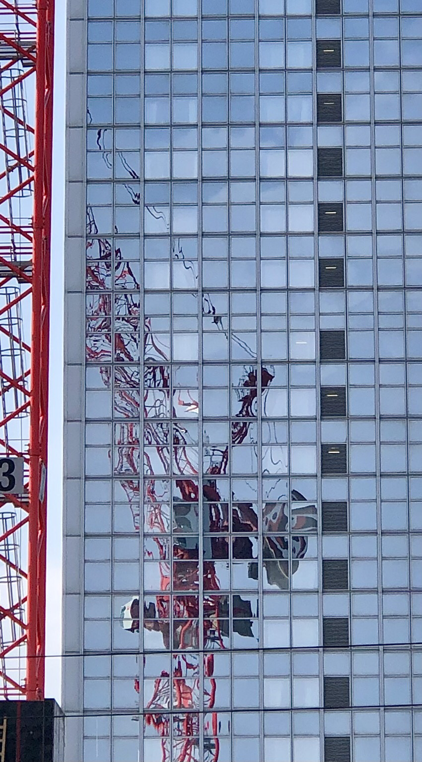Fassade eines Hochhauses mit quadratischer Fenstereinteilung; auf der graublauen Glasfläche sind die verzerrten roten Linien der Spiegelung eines roten Baukrahns zu sehen; links neben Hochhaus ist ein Teil der roten Stahlkonstruktion des Baukrahns zu sehen; rechts in der Glasfassade gibt es eine Reihe dunkler Fensterquadrate, die senkrecht untereinander angeordnet sind.