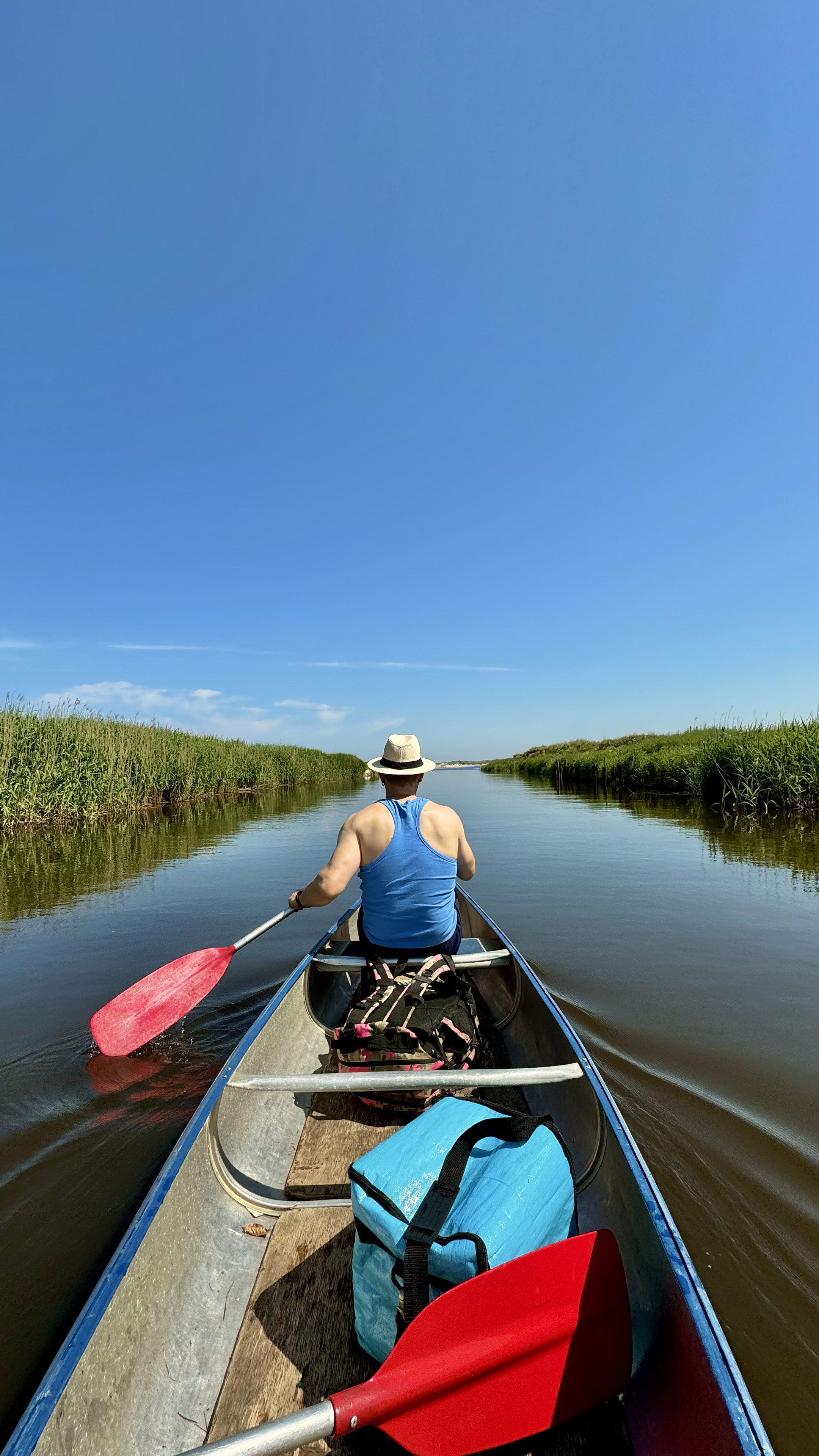 Ein Fluss, aus dem Kanu von hinten fotografiert sieht man vorne einen Ruderer, ein wenig Gepäck im Boot. Links und rechts am Ufer hohes Schilf-Gras unter einem blauen, nahezu wolkenlosen Himmel. Ein Fluss, aus dem Kanu von hinten fotografiert sieht man vorne einen Ruderer, ein wenig Gepäck im Boot. Links und rechts am Ufer hohes Schilf-Gras unter einem blauen, nahezu wolkenlosen Himmel.