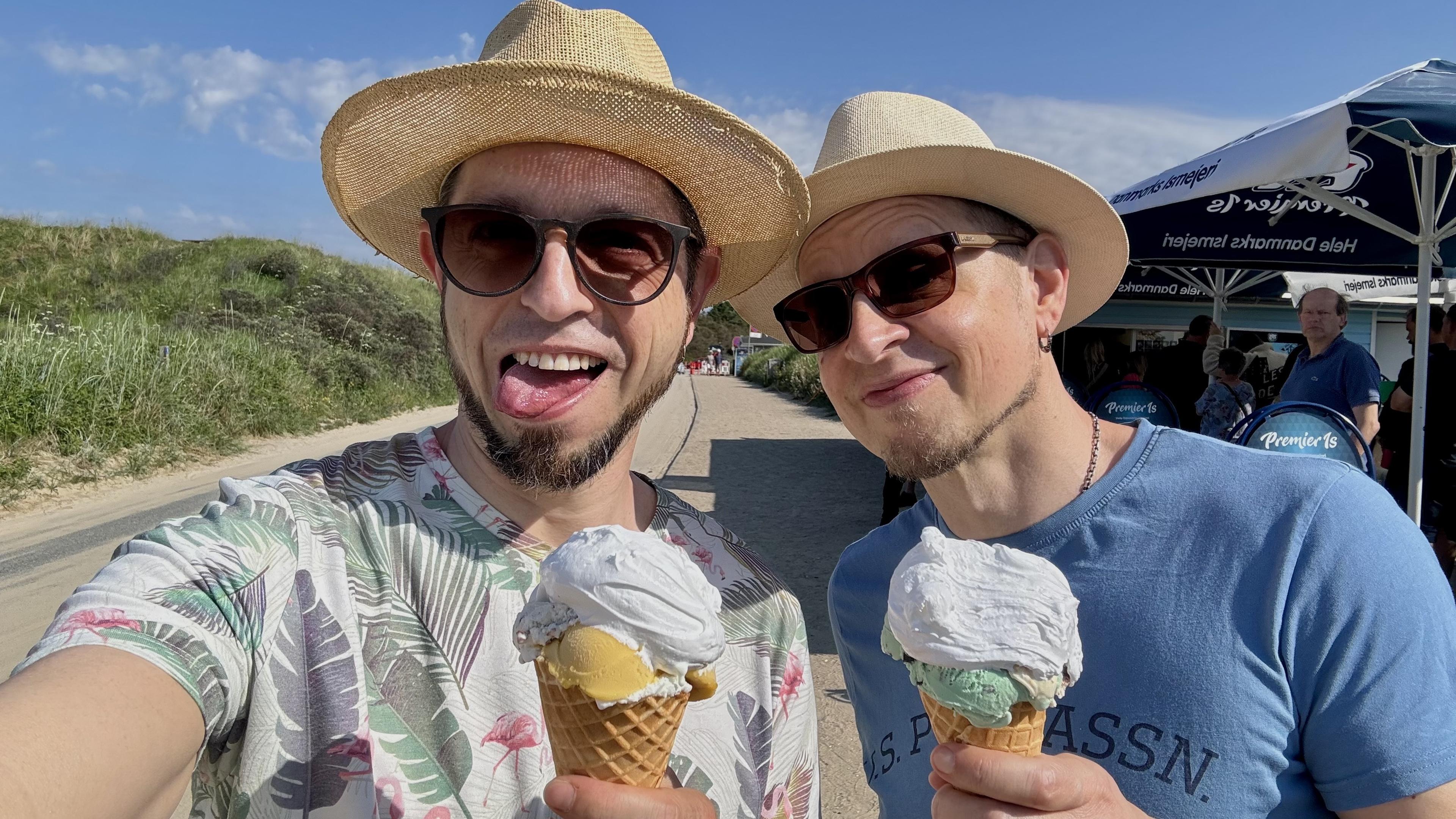 Zwei Männer posieren für ein Selfie auf einem sonnigen Strandweg, beide tragen Strohhüte und Sonnenbrillen. Jeder hält eine Eistüte mit Schlagsahne - eine mit gelbem Eis und die andere mit grünem. Der Hintergrund zeigt Strandgras und einige Wolken. Zwei Männer posieren für ein Selfie auf einem sonnigen Strandweg, beide tragen Strohhüte und Sonnenbrillen. Jeder hält eine Eistüte mit Schlagsahne - eine mit gelbem Eis und die andere mit grünem. Der Hintergrund zeigt Strandgras und einige Wolken.