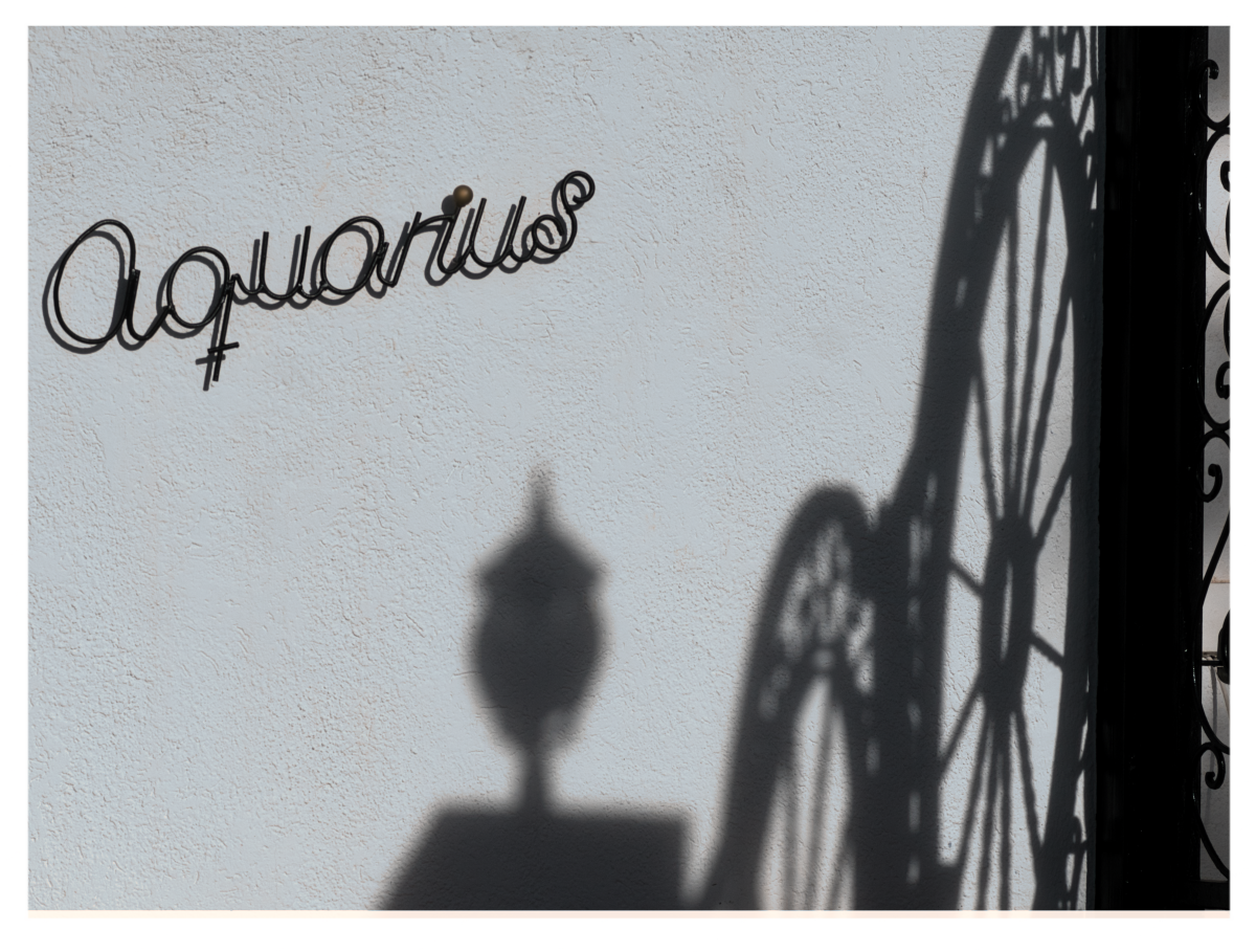 Querformat: Blick auf eine weiss gekalkte Hausmauer, an ihr ist in schmiedeeisernen Buchstaben der Name 'Aquarius' mit etwas Abstand zur Wand befestigt. Rechts die harten Schatten des aufwändig gestalteten Eingangstors in Form von einer Art Radspeichen. Mittig unschärfer eine Eingangslaterne. Mittagswinterlicht.