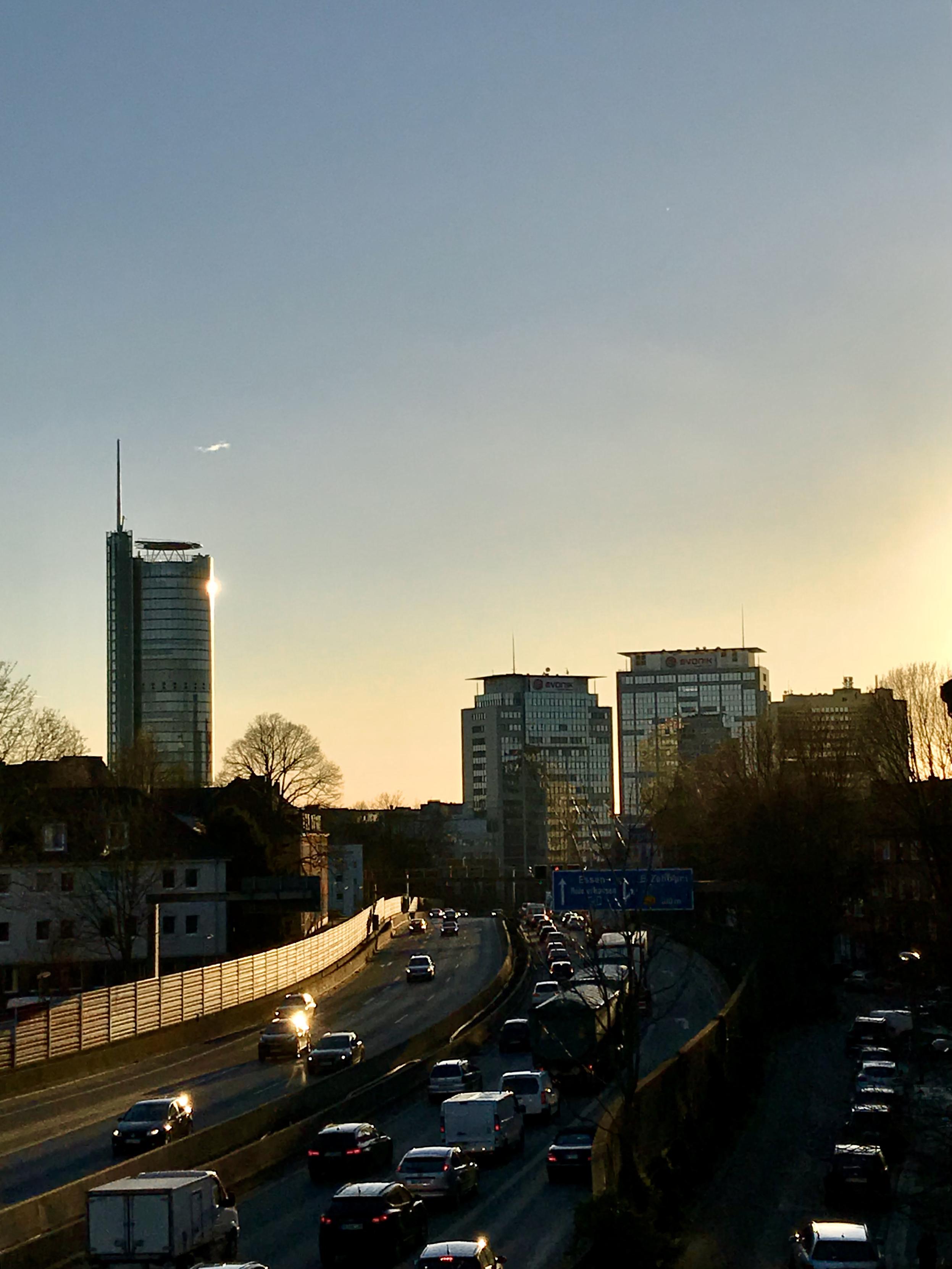 Die tiefstehende Sonne sorg für eine goldige Lichtstimmung. Man sieht Spuren der Autobahn, auf der sich Autos stauen. Im Hintergrund einige Hochhäuser, in deren Glasfronten die Sonnen reflektiert.