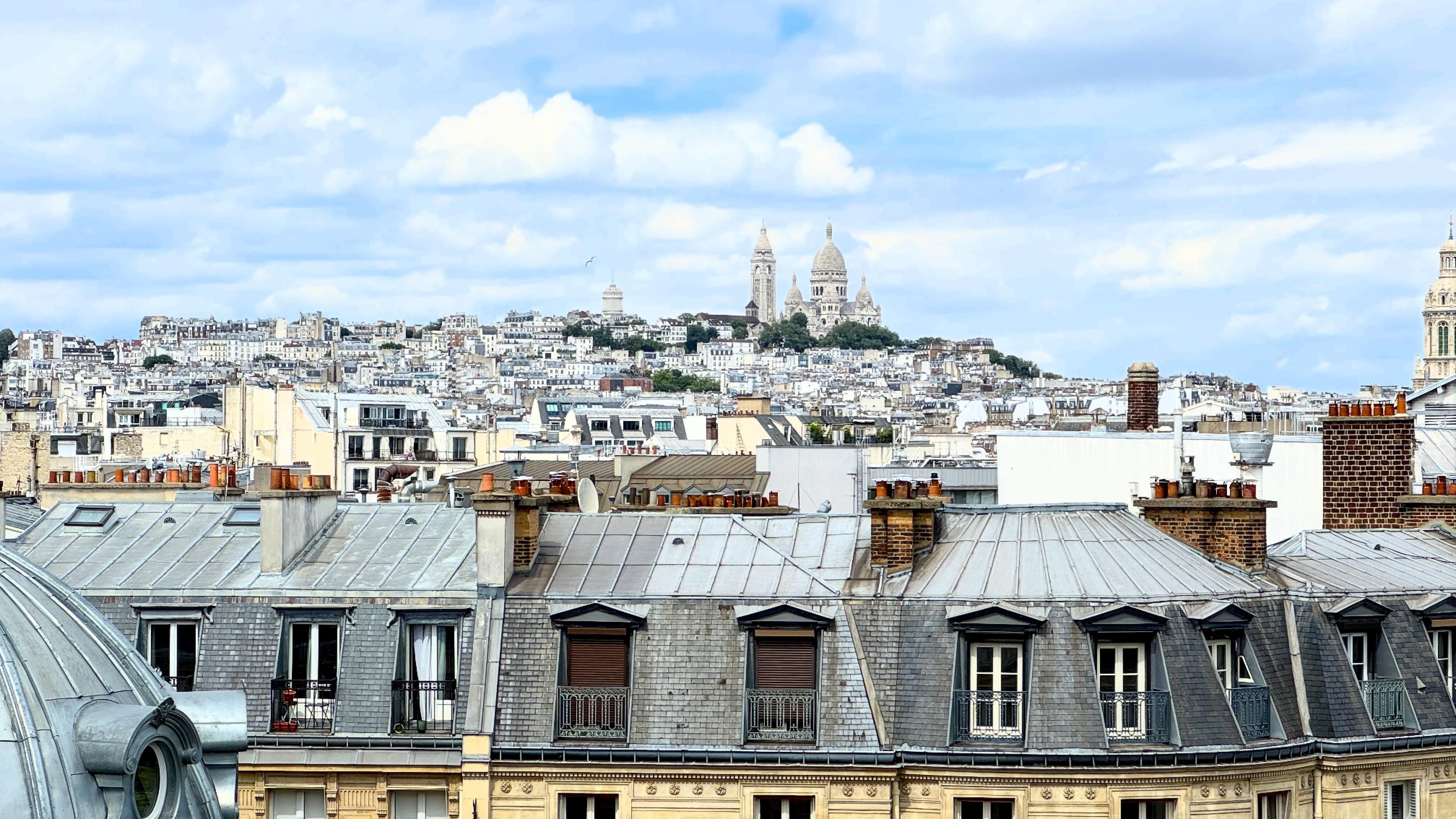 Ein Blick auf Paris mit Dächern im Vordergrund und der Basilika Sacré-Cœur, die prominent auf einem Hügel im Hintergrund unter einem teilweise bewölkten Himmel liegt.