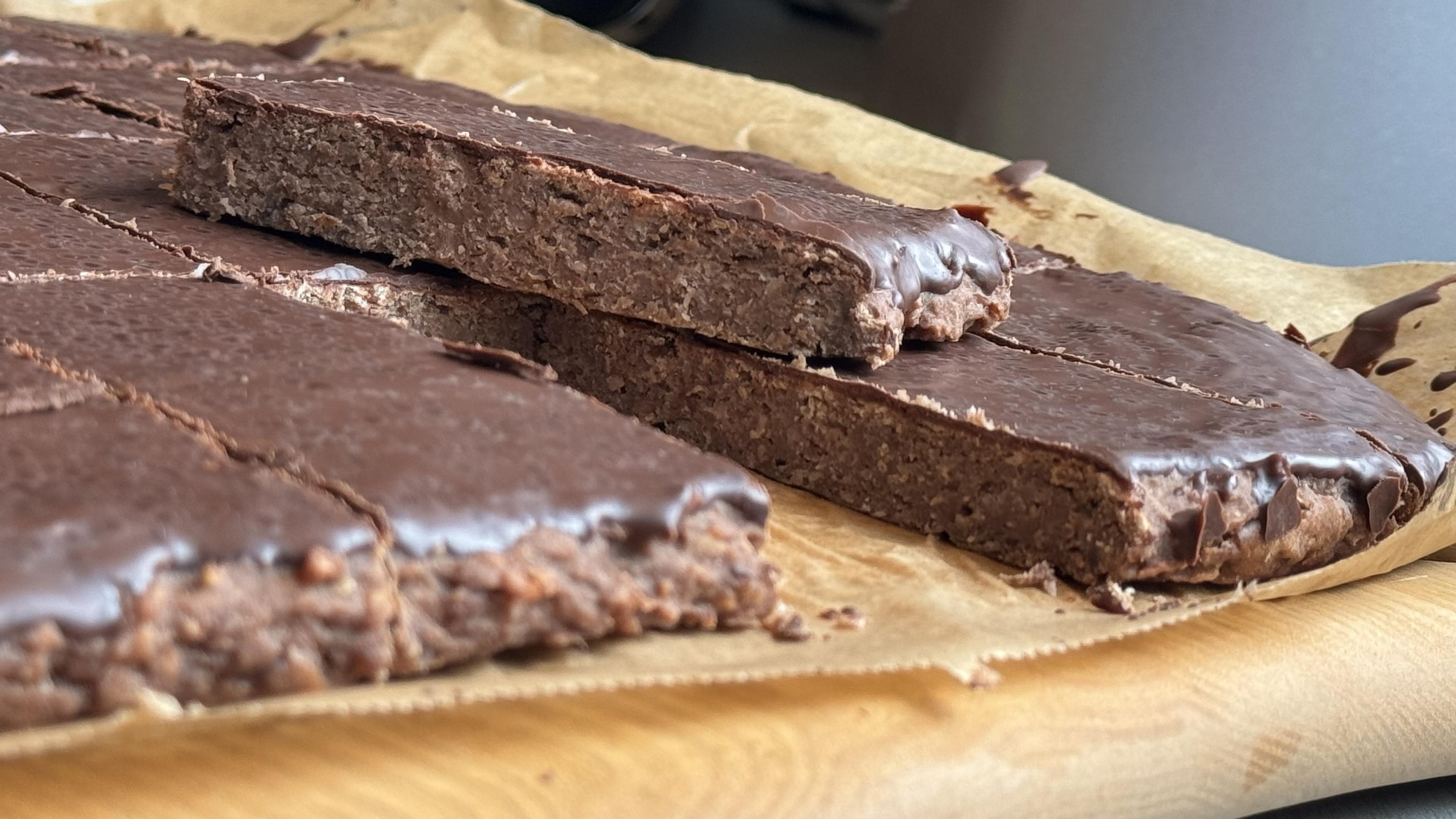 Eine Nahaufnahme von Müsliriegeln, die in rechteckige Stücke geschnitten werden, mit einer glänzenden Schokoladenbeschichtung darauf. Die Stangen sind auf Pergamentpapier auf einer Holzoberfläche angeordnet.