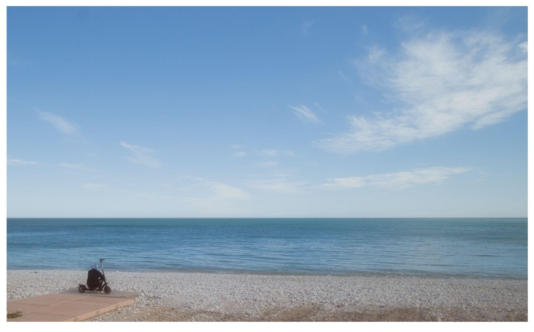 Querformat: heller Himmel mit zerfaserten Wolken über blauem Meer. Links am Ende eines Decks das zum Strand führt, sitzt eine schwarzgekleidete Person mit heller Kapuze auf einem Tretroller und blickt aufs Meer.
