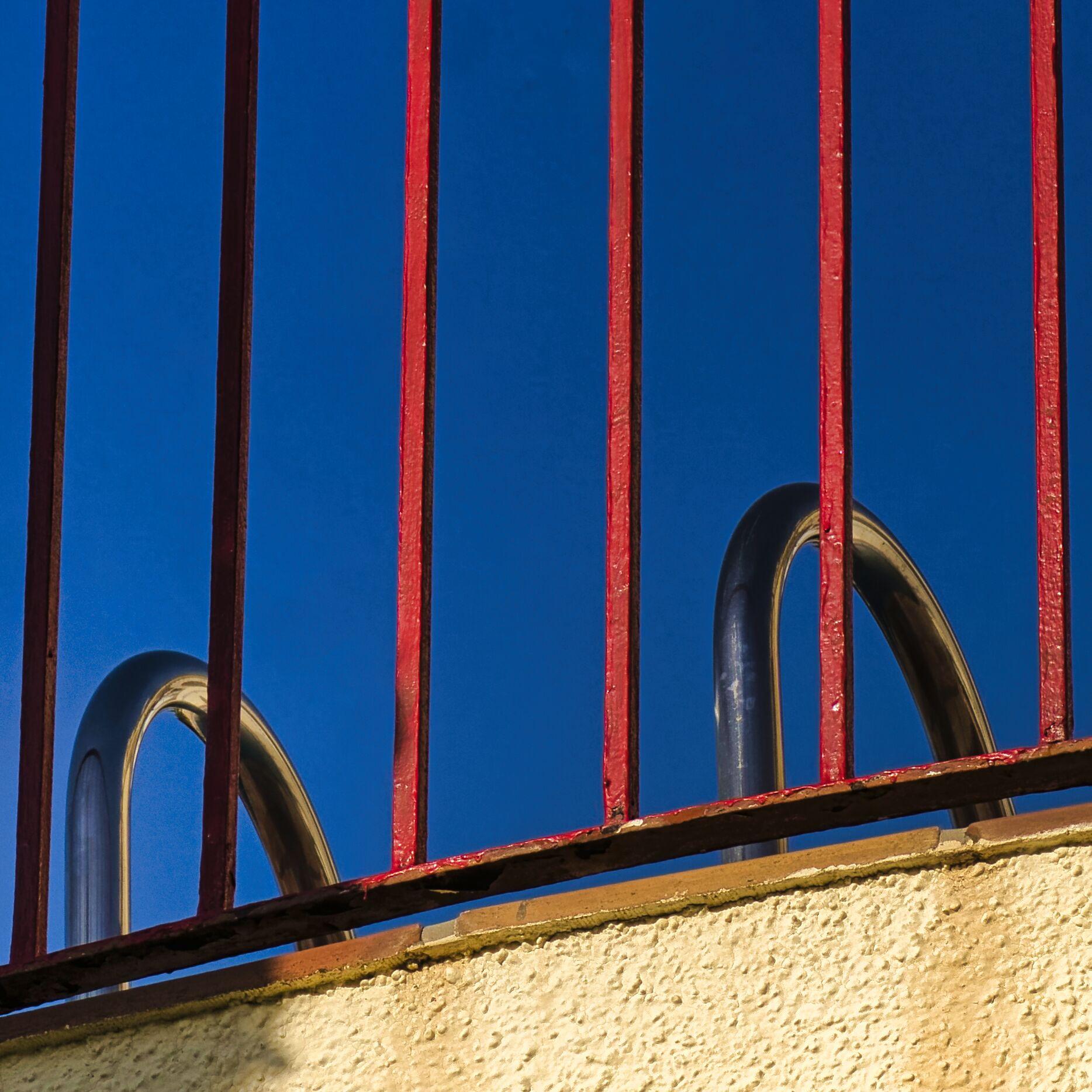 Blick aus der Froschperspektive hinauf zu einem rot gestrichenem Gitter, zwei chromfarbene gebogene Halterungen als Einstigshilfe in einen Pool. Mediterraner tiefblauer Himmel.