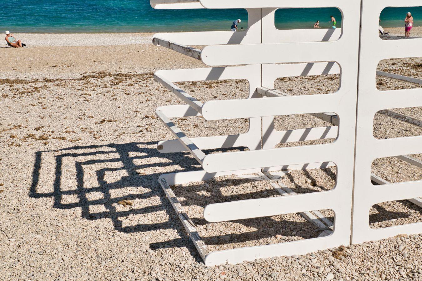An einem Kieselstrand am Meer steht ein weiss gestrichenes Holzgestell, dessen harter Schatten ein sich überlappendes grafisches Muster bildet. Im Hintergrund verschiedene Personen in Badebekleidung.