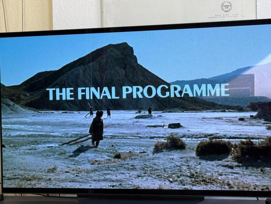 Foto eines Fernsehers, darauf: Eine Wüstenlandschaft, Berge im Hintergrund. Schattenhafte Gestalten scheinen nach rechts zu gehen. Schriftzug „The Final Programme“.